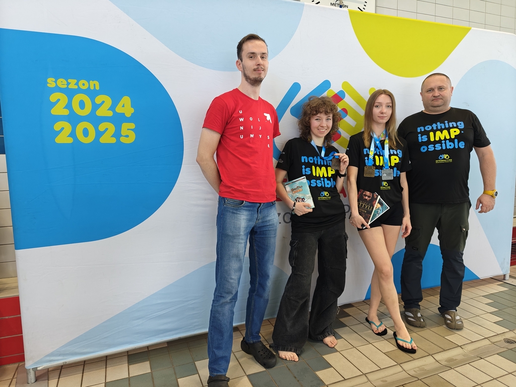 Cztery osoby pozują na tle kolorowego banera z napisem „sezon 2024/2025”. Dwie młode kobiety stoją po środku, ubrane w czarne koszulki z napisem „nothing is IMPossible”; trzymają medale i książki. Towarzyszą im dwaj mężczyźni – jeden w czerwonej koszulce po lewej i drugi w czarnej po prawej. Wszyscy stoją na basenowej posadzce, sugerując udział w wydarzeniu sportowym lub konkursie.